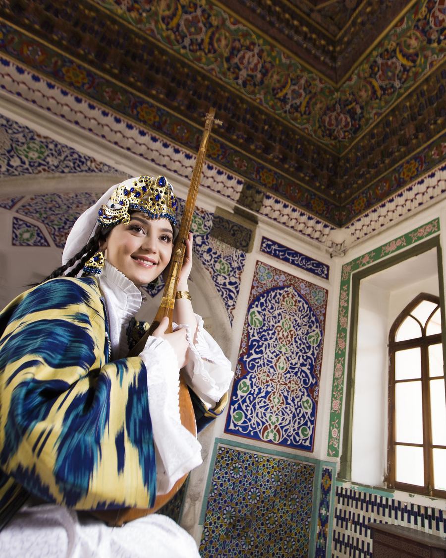 A young woman in an ikat chapan and jewelled headdress playing a dutar beneath painted Uzbek tilework